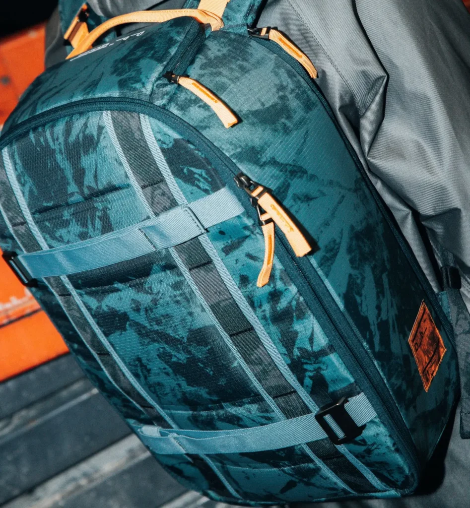 A teal-green backpack with a subtle camo-like pattern, featuring multiple zip compartments with orange pull tabs. The front panel includes horizontal compression straps and a central vertical webbing system for modular attachments. The fabric appears water-resistant, paired with a contrasting gray side panel, giving it a rugged, outdoor-ready aesthetic with functional detailing.