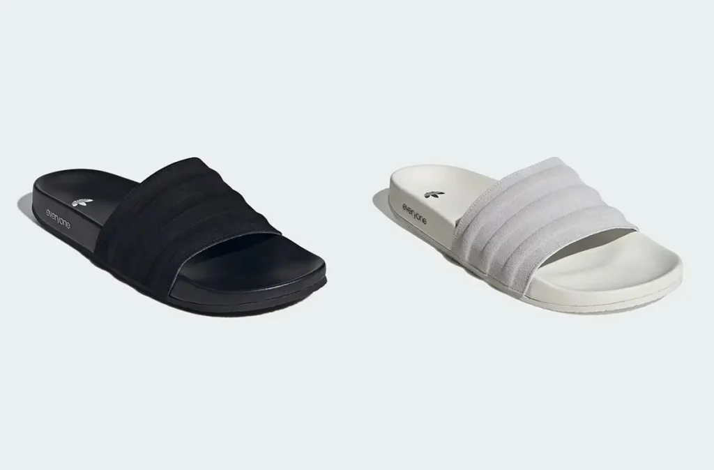A side-by-side product shot of two Adidas slide sandals presented against a clean, neutral background. On the left is an all-black version, while on the right is a light grey/cream variant. Both feature a wide, ribbed strap across the upper and a smooth contoured footbed for comfort. Subtle branding appears along the side of the sole with the word “everyone,” accompanied by a small Adidas logo near the heel. The minimalist color palette and simple silhouette emphasize versatility and understated design