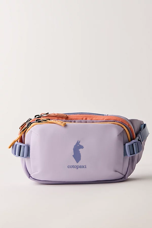 Compact Cotopaxi crossbody bag in light lavender with orange and yellow zipper accents, featuring a front logo print, multiple zip compartments, and an adjustable strap with side buckles