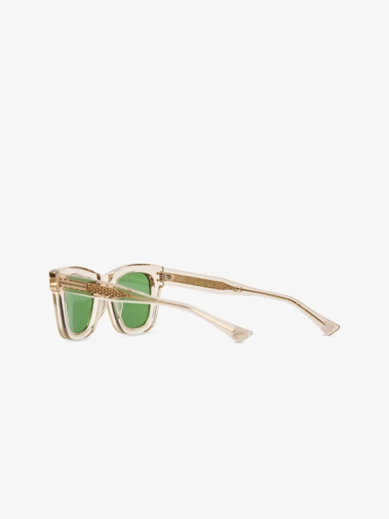 Transparent champagne acetate sunglasses shown in side profile, featuring green lenses, exposed gold-tone hinge construction, engraved metal core temples, and slim tapered arms with refined minimal detailing