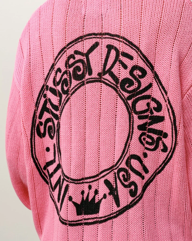 Close-up detail of a pink BUANA ribbed knit sweater showcasing a textured vertical stitch pattern and a bold black circular logo with stylized script lettering and a crown emblem at the bottom