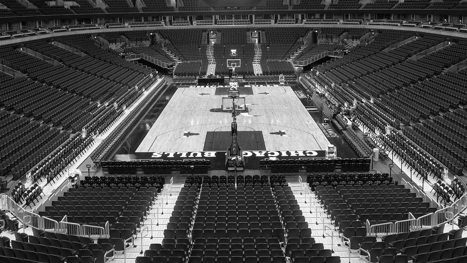 Black-and-white wide-angle view of an empty Chicago Bulls basketball arena, showing the full court centered beneath the scoreboard, surrounded by neatly arranged rows of seats and steep tiers of stands extending upward in symmetrical formation