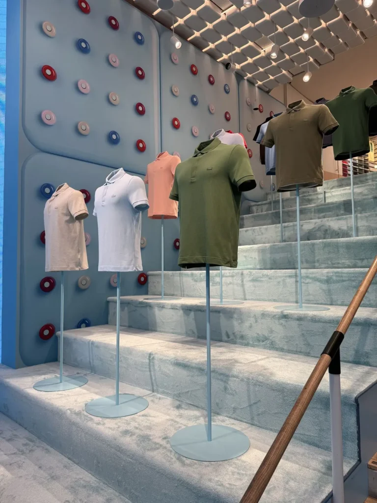 Lacoste Polo Factory Paris display featuring classic polo shirts in multiple colors presented on elevated stands within a pastel-toned, Atomic Age-inspired exhibition space