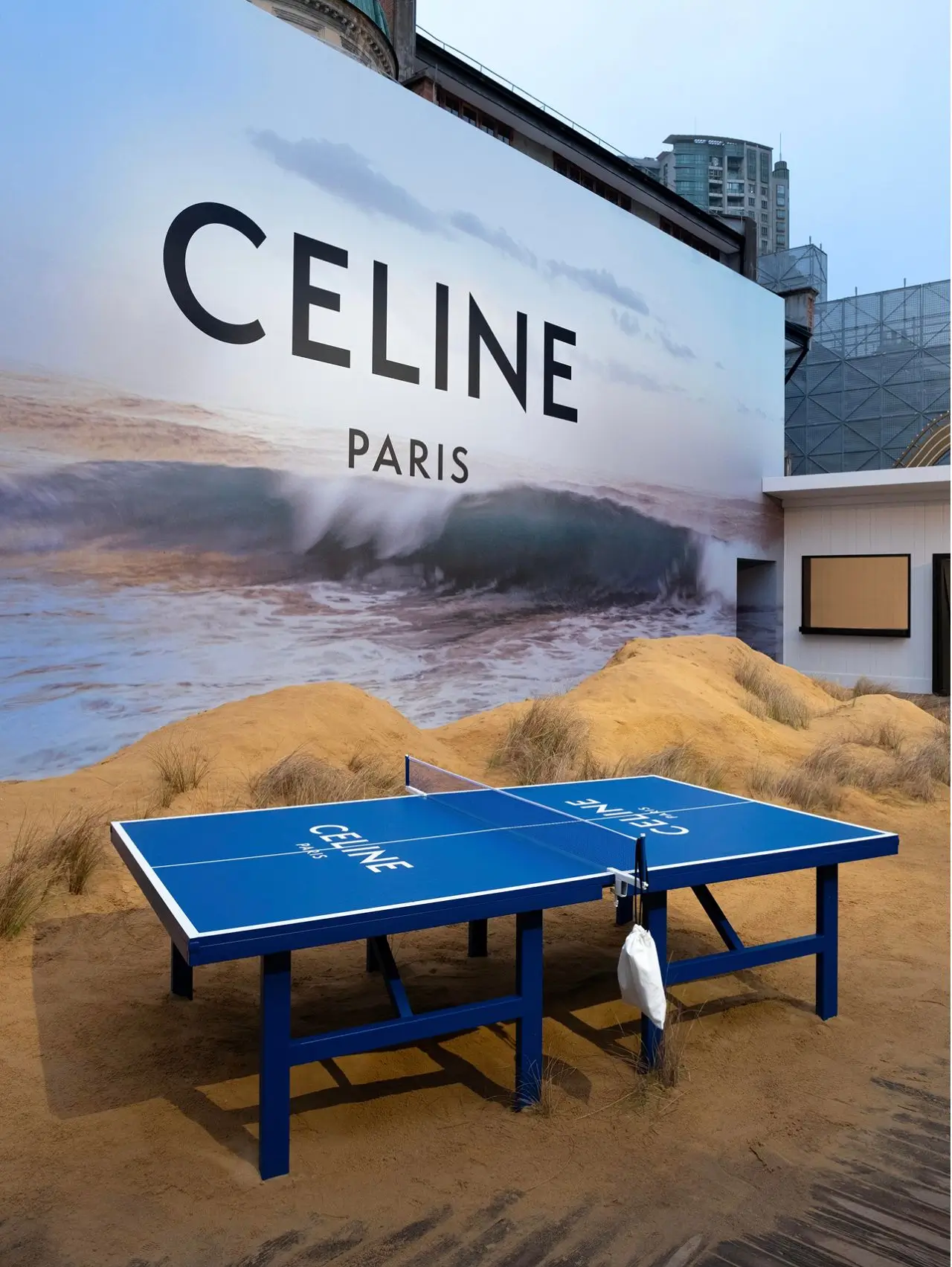 Celine-branded blue ping pong table set in sand with a large ocean-wave mural and “CELINE PARIS” signage on a building facade in the background