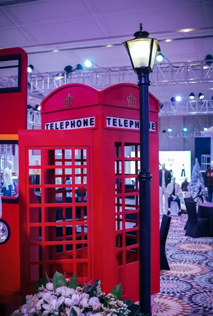 A bright red British telephone booth display stands indoors beside a vintage-style black streetlamp, surrounded by floral arrangements and exhibition lighting trusses, evoking a London streetscape within a fashion showroom setting