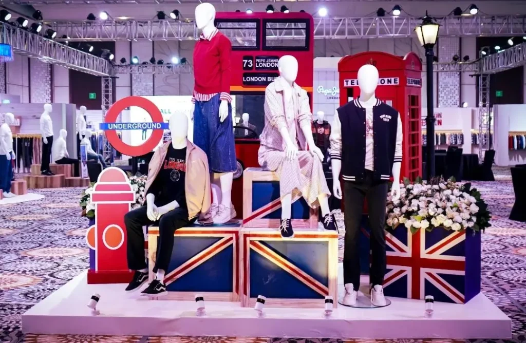 A retail presentation featuring mannequins dressed in casual apparel is staged against a London-themed backdrop, including a red double-decker bus, Underground sign, telephone booths, and Union Jack platforms, all arranged within a large indoor exhibition space