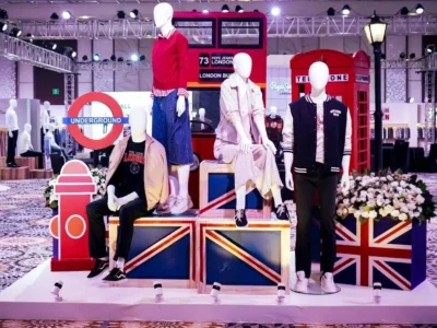 A retail presentation featuring mannequins dressed in casual apparel is staged against a London-themed backdrop, including a red double-decker bus, Underground sign, telephone booths, and Union Jack platforms, all arranged within a large indoor exhibition space