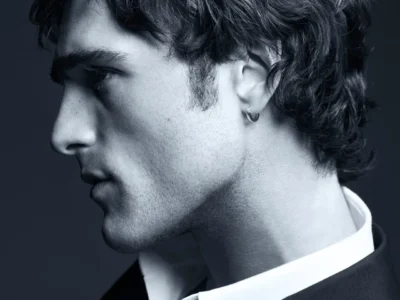 A black-and-white close-up portrait of Jacob Elordi, his head turned slightly downward. He has tousled, medium-length hair and light stubble along his jawline, with a small hoop earring in his ear. The lighting is soft yet directional, emphasizing the contours of his cheekbones, nose, and neck. He wears a crisp collared shirt beneath a tailored jacket, giving a refined, understated look. The overall mood is contemplative and minimal, with a clean studio background enhancing the sculptural quality of his features