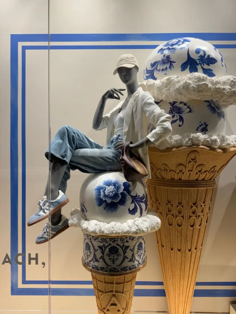 Store window display featuring a seated mannequin styled in a white cap, white top, blazer, and light blue jeans, paired with casual sneakers and a small shoulder bag. The mannequin rests against oversized decorative elements resembling ice cream cones topped with blue-and-white porcelain-style spheres and textured “cream,” set against a clean backdrop with blue graphic lines