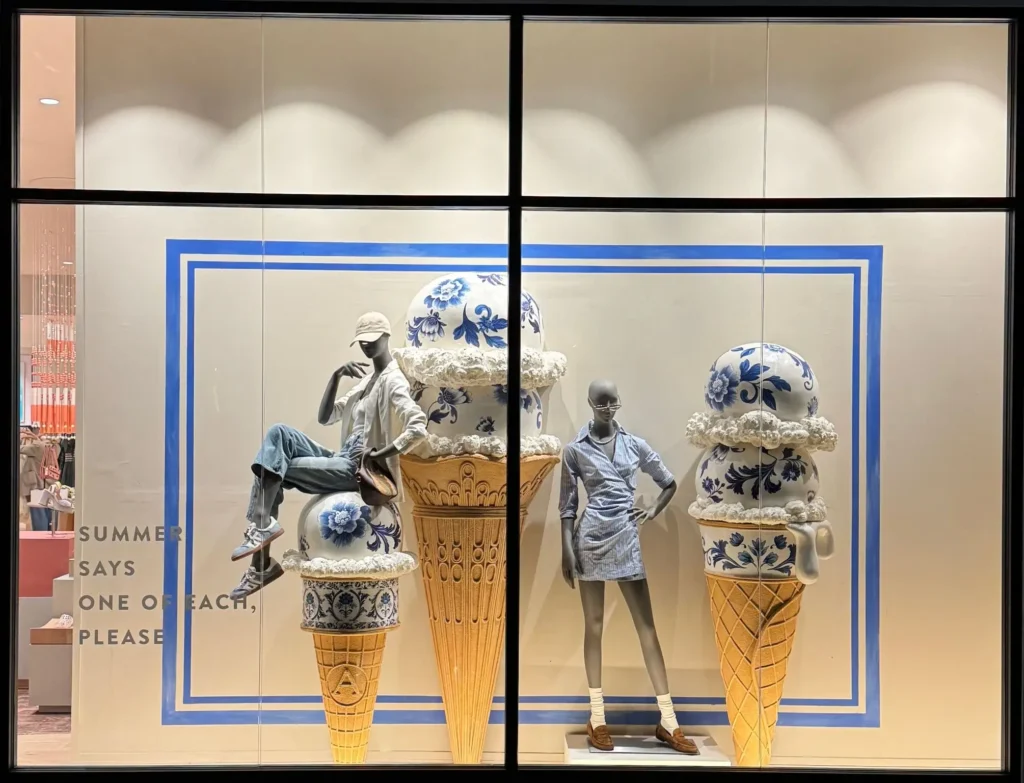 Wide view of a retail storefront window display featuring two mannequins styled in casual summer outfits—one seated and one standing—posed among oversized ice cream cone sculptures topped with blue-and-white porcelain-style spheres and textured “cream.” The scene is framed by blue graphic lines, with text on the glass reading “SUMMER SAYS ONE OF EACH, PLEASE,” view across the display