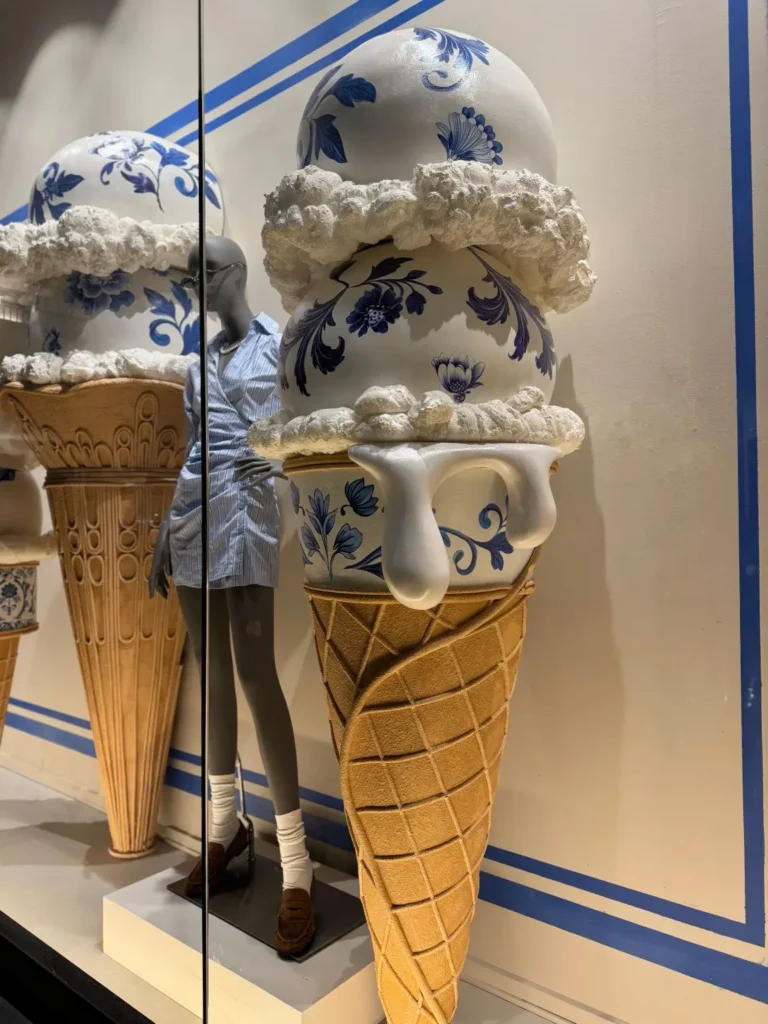 Close-up of a storefront display featuring an oversized ice cream cone sculpture with a waffle-textured base and a porcelain-style scoop decorated with blue floral patterns, topped with textured “cream” layers and dripping white detail. A mannequin in a light blue shirt dress stands beside it, partially reflected in the glass, against a clean backdrop with blue graphic lines