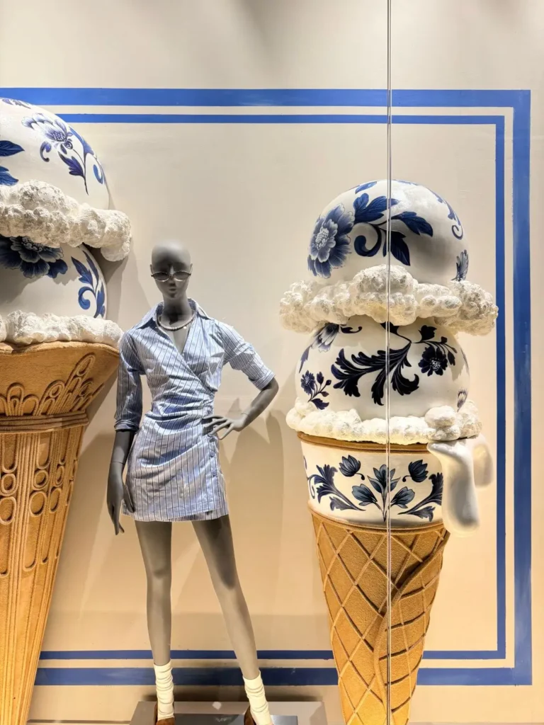 Store window display featuring a standing mannequin styled in a light blue striped shirt dress with white socks and brown loafers, posed beside oversized ice cream cone sculptures topped with blue-and-white porcelain-style spheres and textured “cream.” The scene is set against a clean backdrop framed by blue graphic lines, with reflections view on the glass