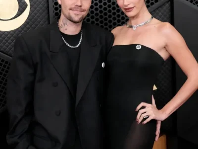 "Justin Bieber and Hailey Bieber posing together on the red carpet at the Grammy Awards. Justin wears a sleek black double-breasted suit with a chain necklace, while Hailey stuns in a strapless black gown with a silver heart choker necklace. The couple stands confidently against a dark geometric backdrop with the Grammy trophy view
