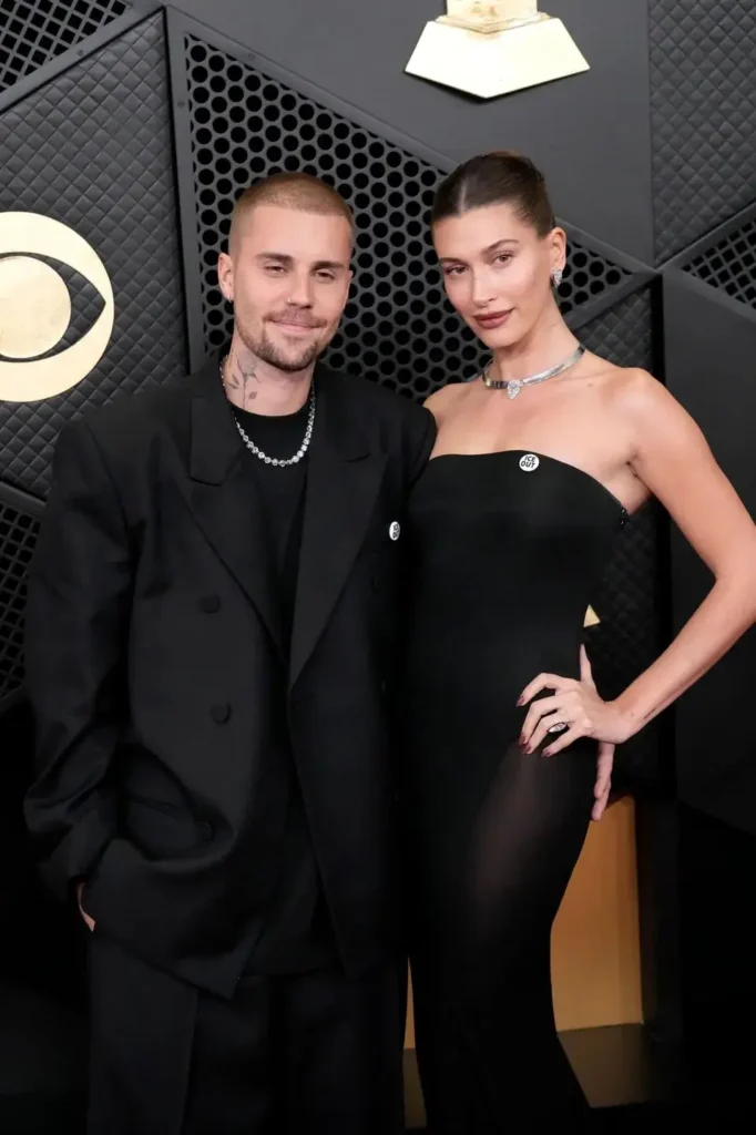 "Justin Bieber and Hailey Bieber posing together on the red carpet at the Grammy Awards. Justin wears a sleek black double-breasted suit with a chain necklace, while Hailey stuns in a strapless black gown with a silver heart choker necklace. The couple stands confidently against a dark geometric backdrop with the Grammy trophy view