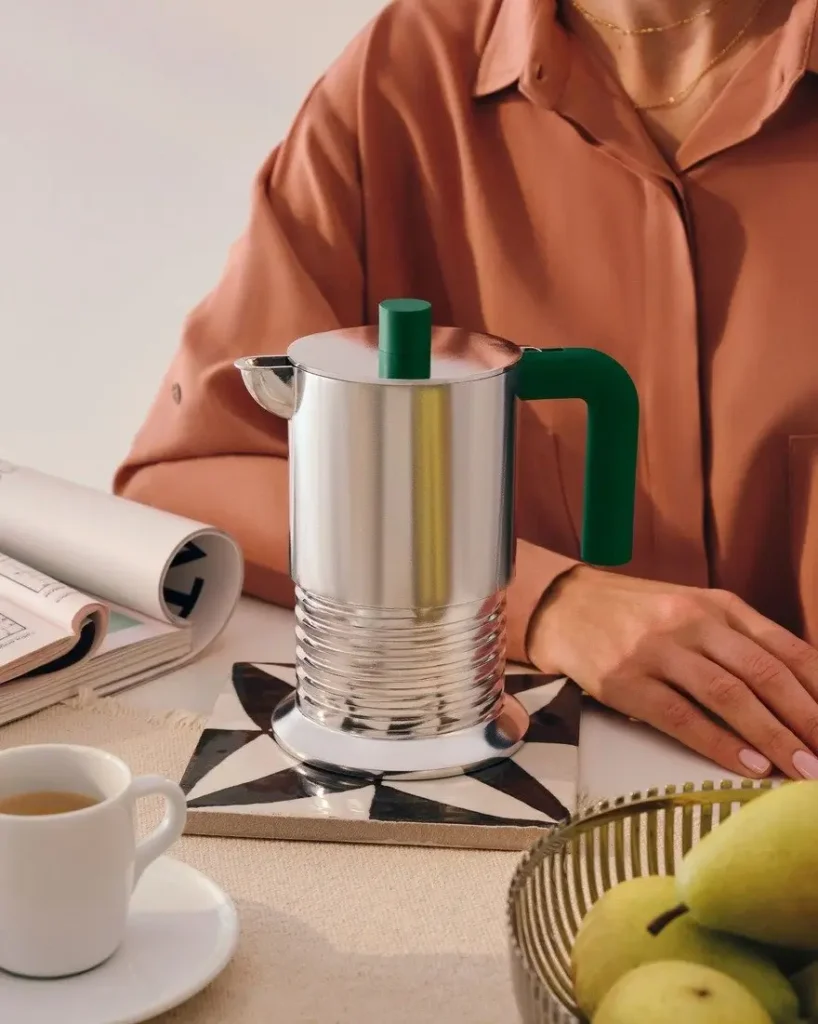 Alessi Vite espresso maker by Philippe Malouin with polished aluminum body and green handle placed on a patterned trivet beside a coffee cup and fruit bowl in a styled kitchen setting