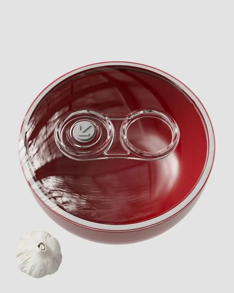 Top-down product shot of the vibrant red B!POD DRO!D charging base from the food vacuum system. The glossy circular dock features a transparent central docking area with two precision ports and a visible vacuum valve, accented by a clean silver rim. A garlic bulb provides scale, emphasizing the premium Italian design and compact footprint. Hi-tech kitchen storage solution, 2026