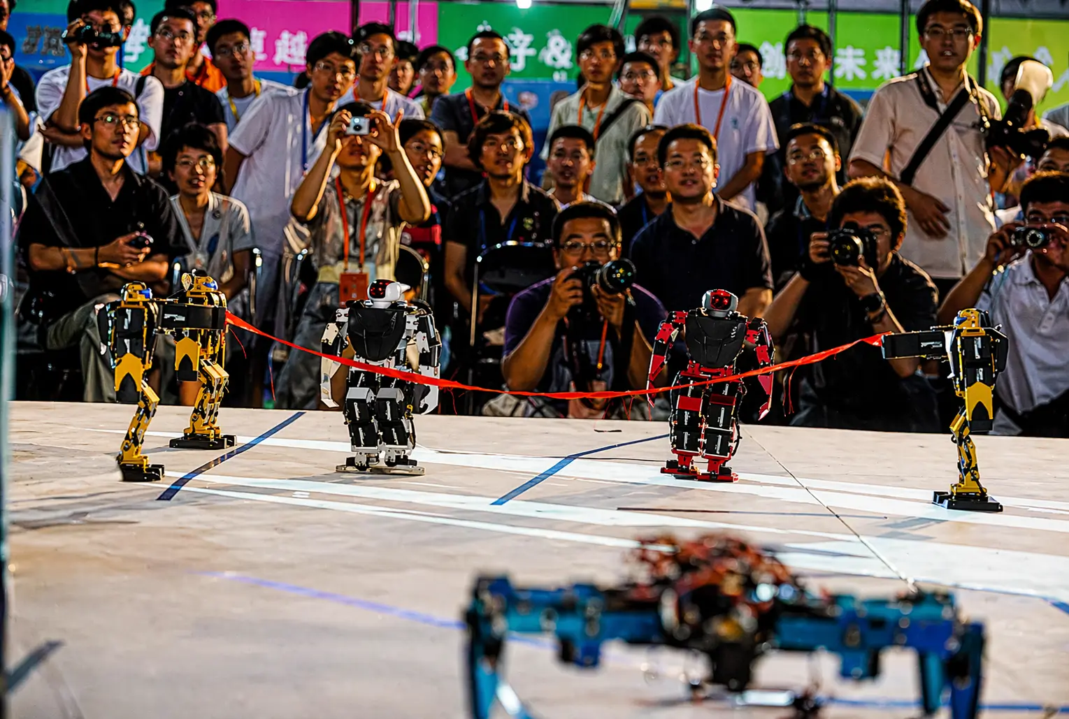 Small humanoid robots race toward a finish line with a red ribbon as a crowd of spectators and photographers watches closely at a robotics event