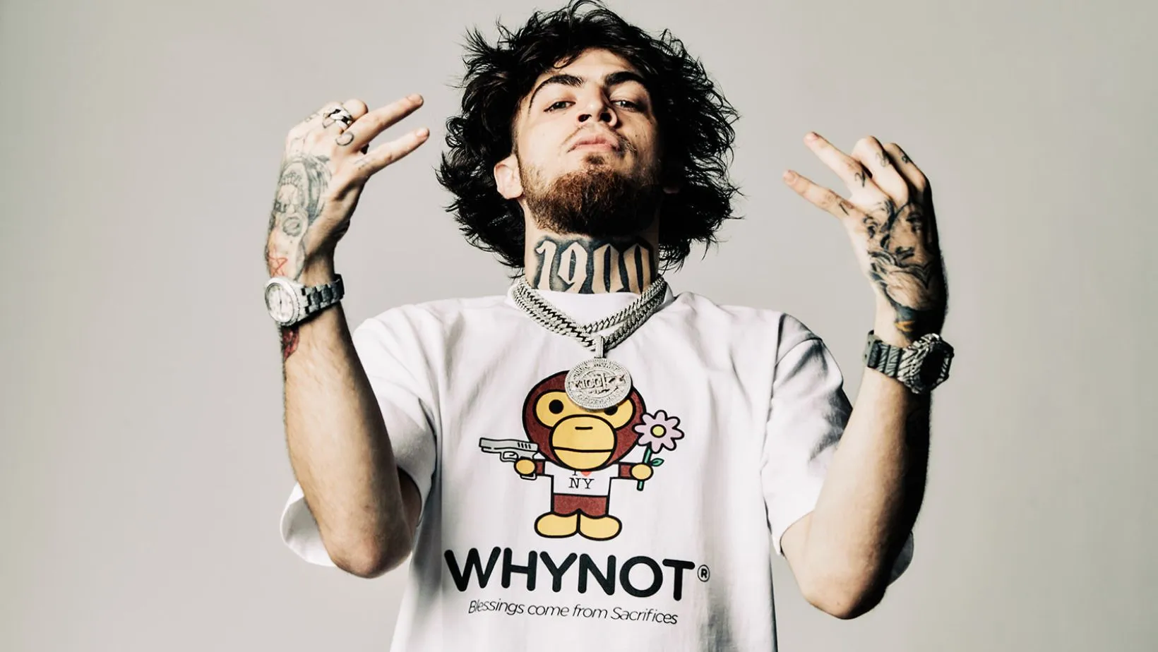 Portrait of 1900Rugrat wearing a graphic “WHYNOT” T-shirt and layered chains, raising both hands to display tattoos against a clean studio background