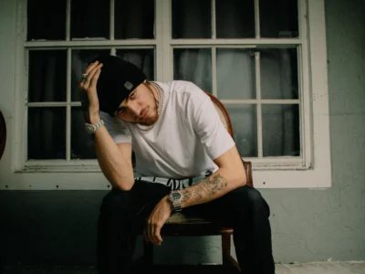Portrait of 1900Rugrat seated on a wooden chair in front of a window at night, wearing a white T-shirt, black beanie, and shoe, conveying the stripped, introspective mood of “Plane Jane”