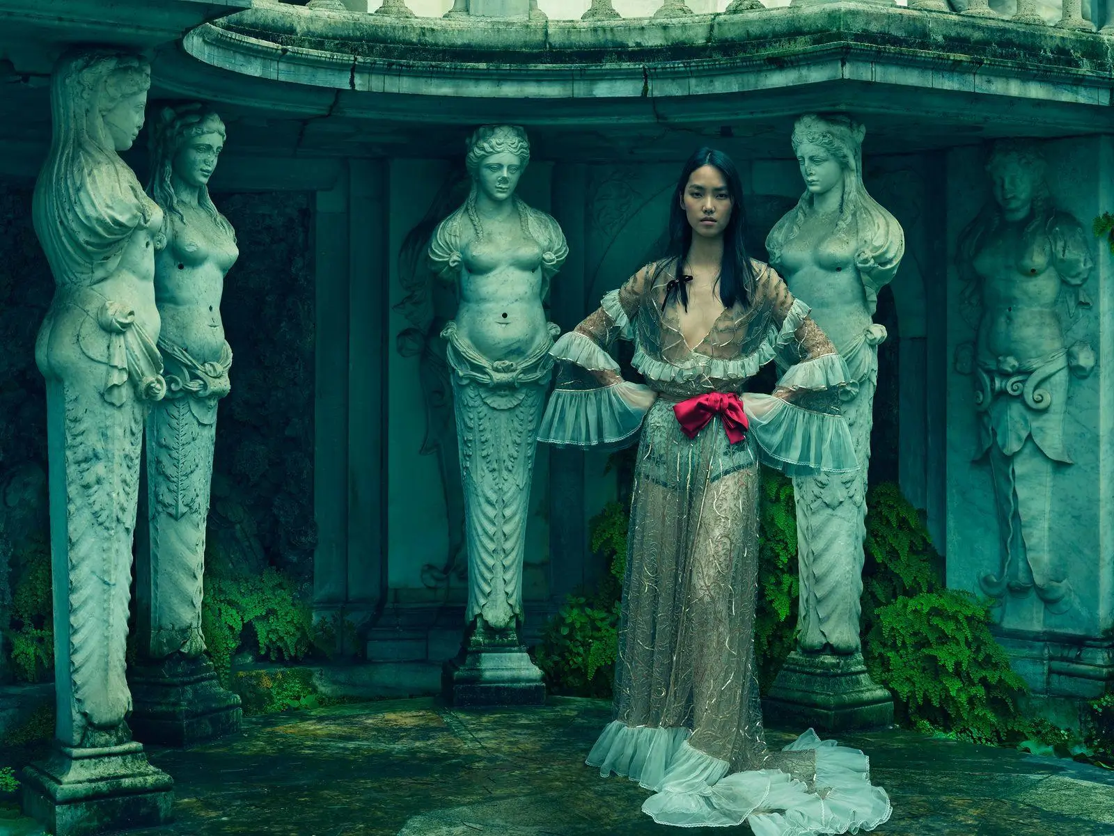 A fashion portrait set in a classical stone alcove, where a model in a sheer, flowing gown with ruffled sleeves and a red ribbon at the waist stands centered among sculpted female figures; the cool green tones, damp stone floor, and surrounding foliage create a moody, antiquity-inspired atmosphere