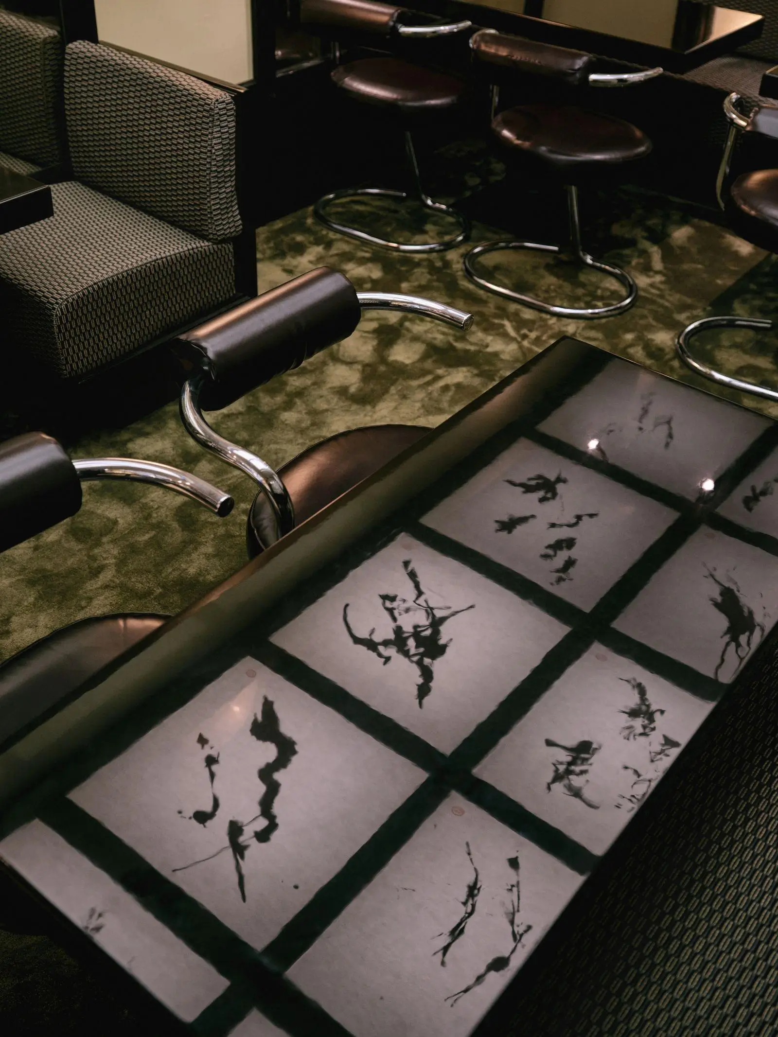 Overhead view of a glossy black table inset with a grid of panels featuring abstract ink-like artwork, surrounded by chrome-framed bar stools and patterned seating, set against a textured green floor in a dimly lit, retro-modern interior