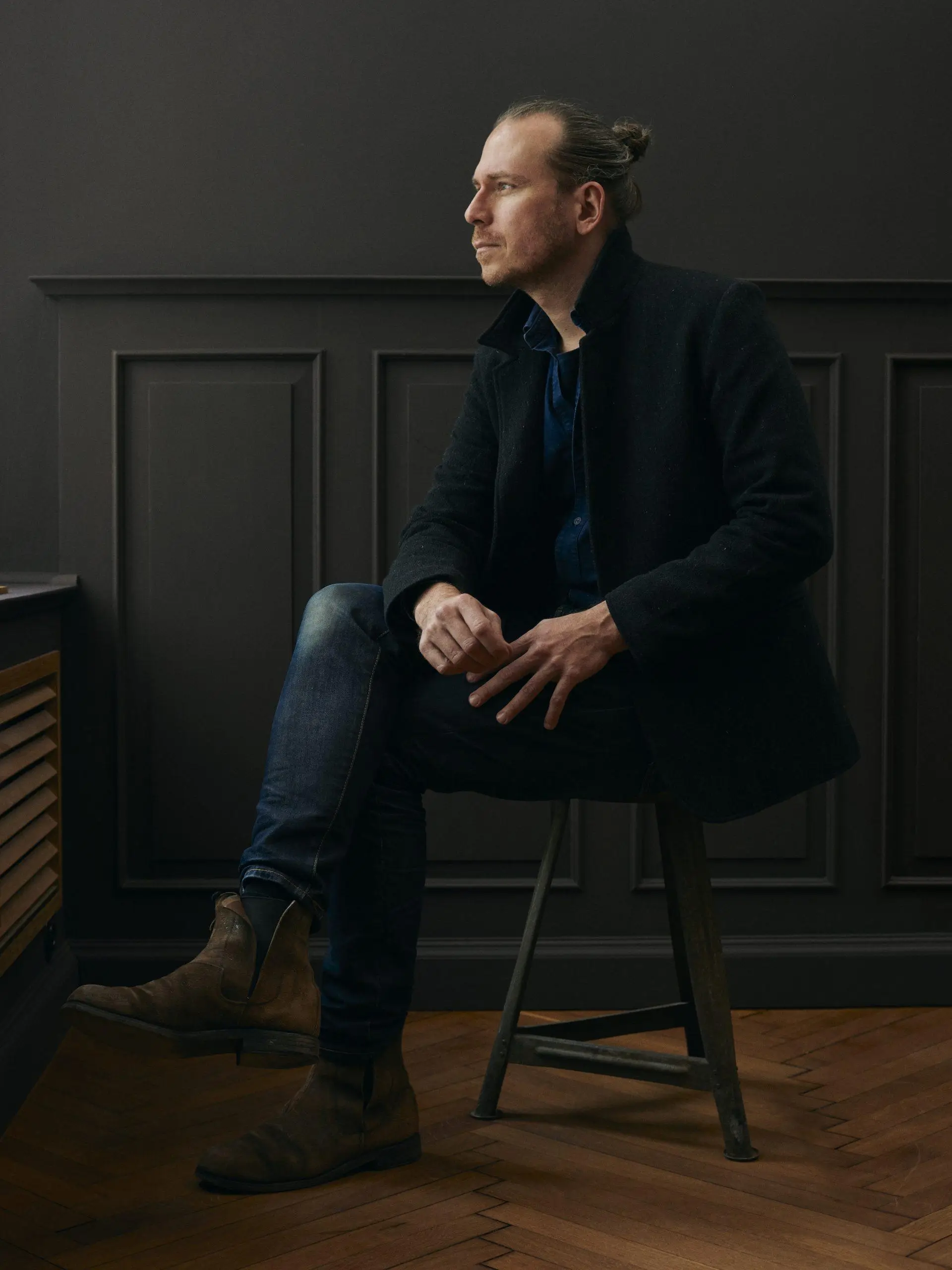 Frank Leder seated on a wooden stool in a dark-paneled room, wearing a black coat, dark denim, and brown leather boots, looking sideways in a contemplative pose