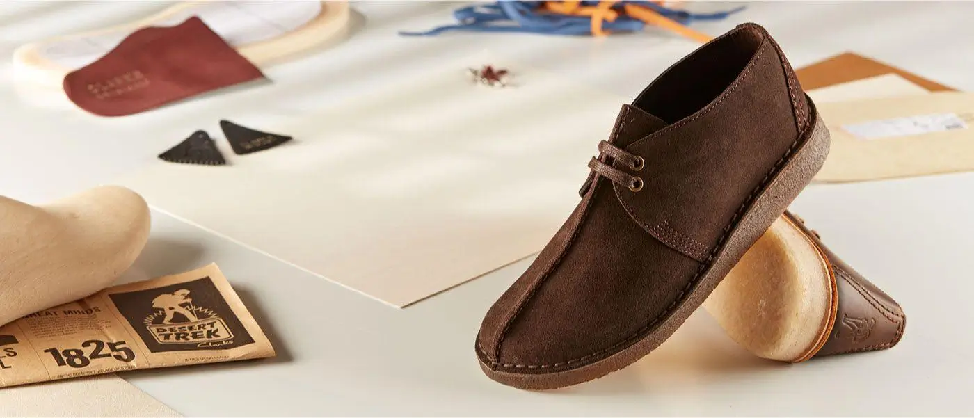 Brown leather Desert Trek–style shoe with a moc-toe construction and two-eye lace system, set against a workshop backdrop with tools, leather pieces, and a crepe sole displayed alongside