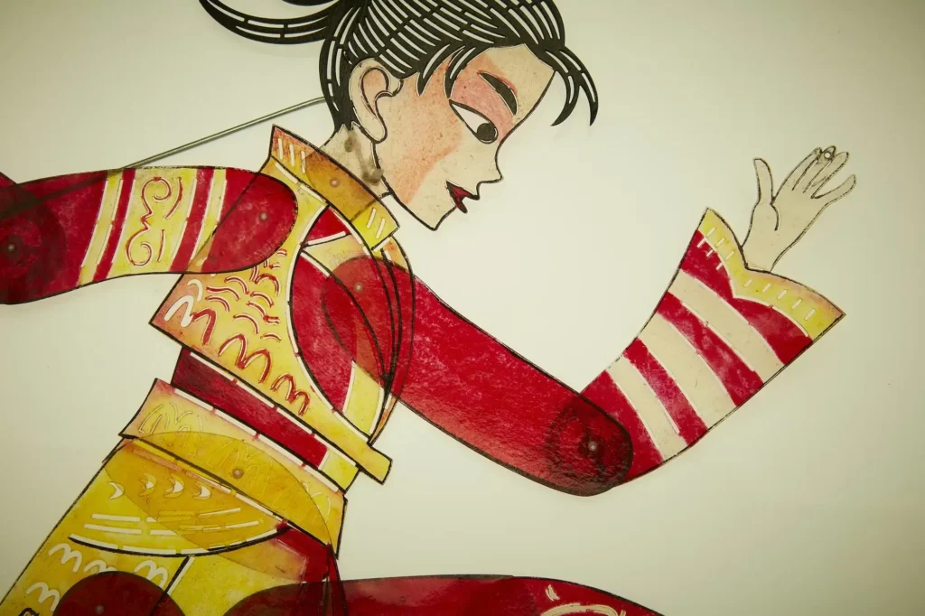 A stylized illustration of a woman in profile, rendered with delicate linework and warm, saturated tones. She wears a red-and-gold patterned garment with striped sleeves and ornamental detailing, while her black hair is swept into a neat bun. Her raised hand, softly poised fingers, and calm expression give the composition a graceful, theatrical sense of movement against the plain background