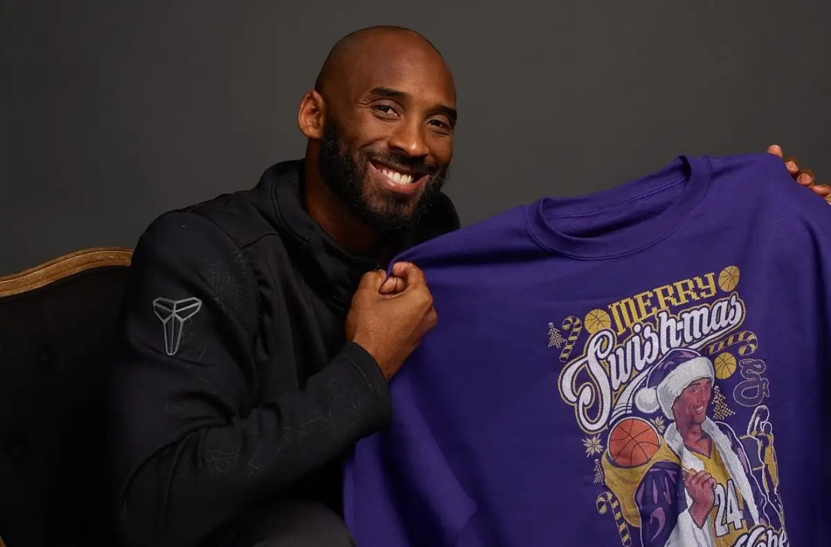 Smiling man seated indoors holding up a purple graphic T-shirt reading “Merry Swishmas,” featuring a festive basketball-themed illustration, against a neutral studio background