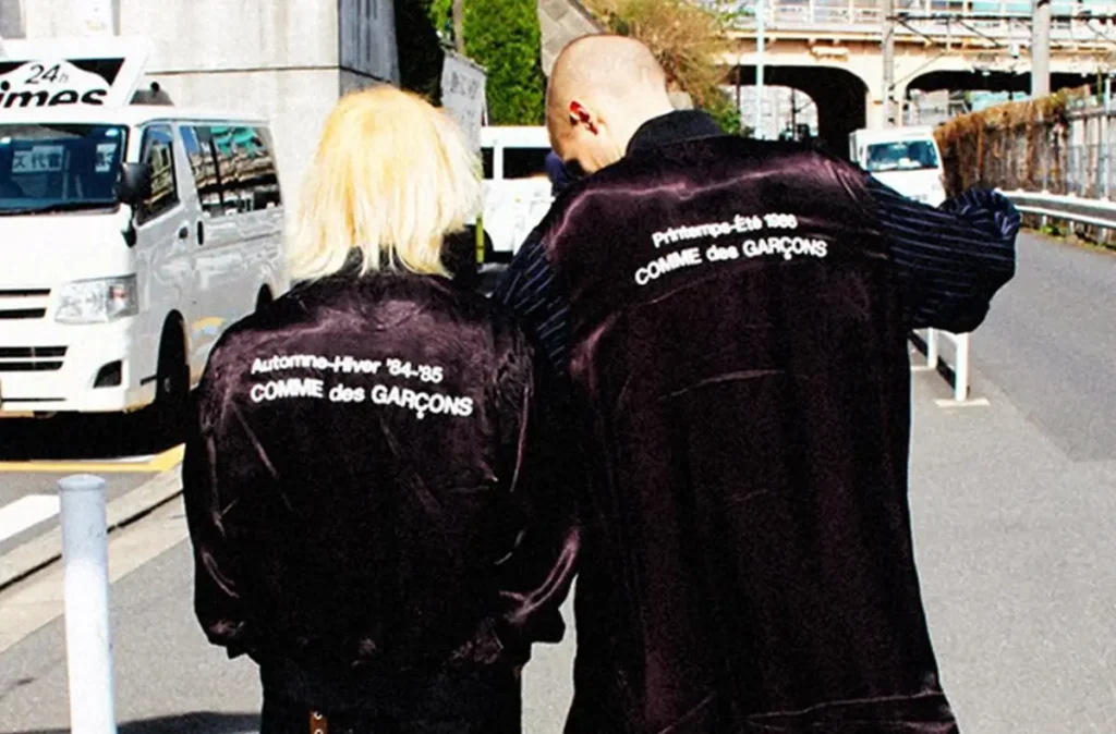 Two figures walking in the city wearing satin Comme des Garçons jackets with archival text on the back, highlighting seasonal references and minimalist typography branding