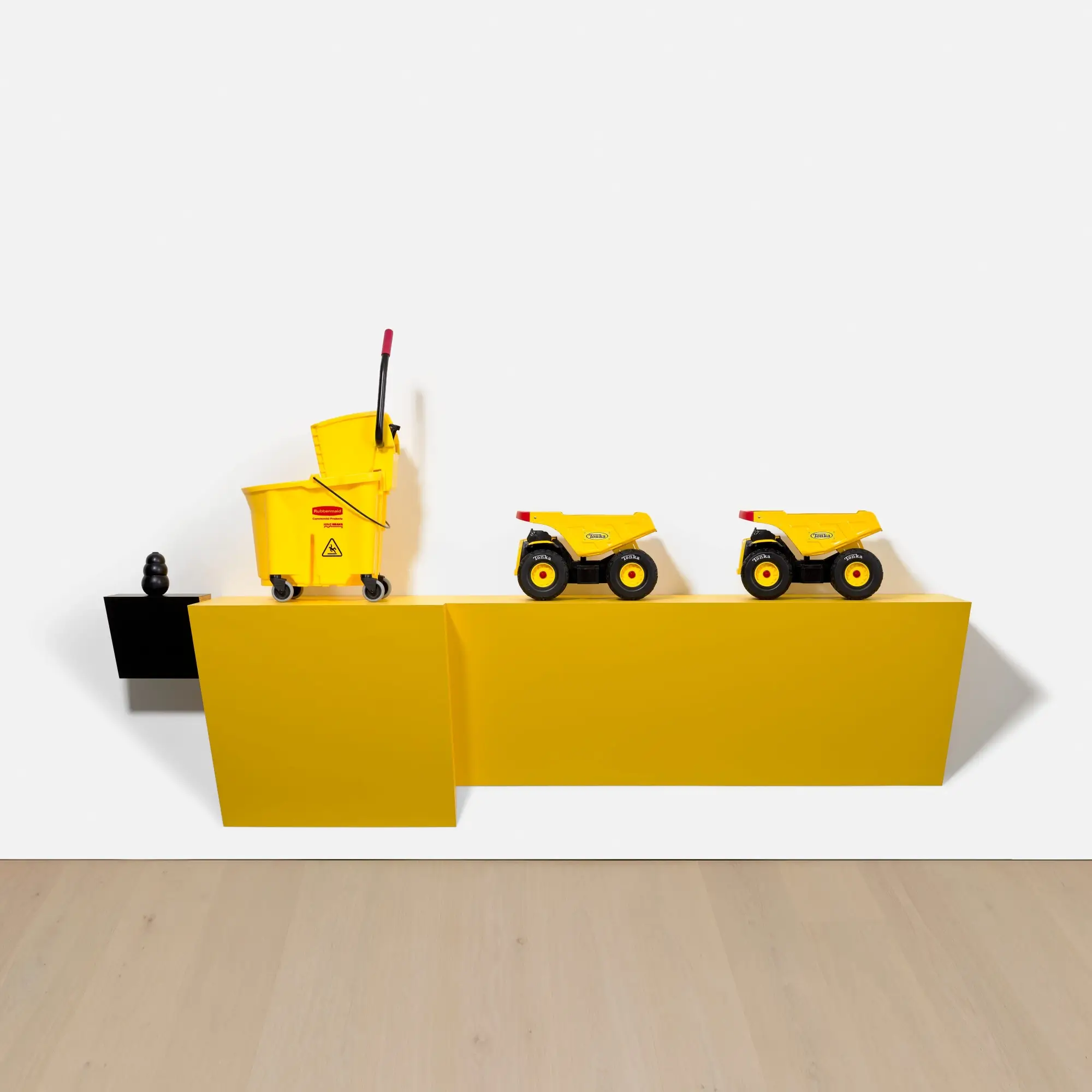 Haim Steinbach installation featuring a bright yellow wall-mounted shelf displaying a Rubbermaid cleaning cart and two yellow toy dump trucks, arranged against a white gallery wall with a small black sculptural object at the far left
