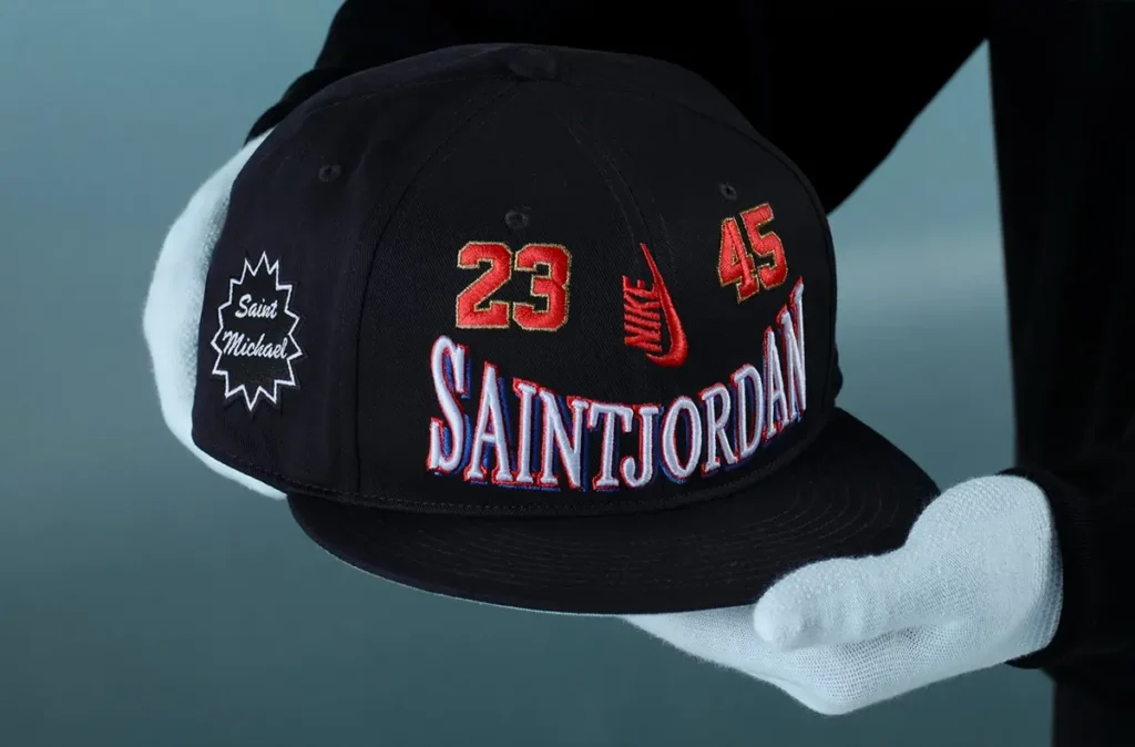Black SAINT Mxxxxxx x Jordan Brand snapback cap held by gloved hands, featuring embroidered “SAINTJORDAN” lettering across the front, red “23” and “45” numerals, a Nike Swoosh detail, and a side patch reading “Saint Michael,” set against a muted grey backdrop