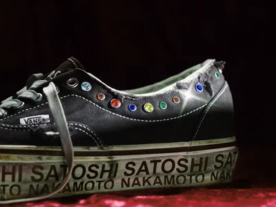 Man walking past a gray exterior wall with large “GUERILLA ATELIER” lettering, framed by a leaning tree and overhead industrial lights, with a parked car partially view in the foreground