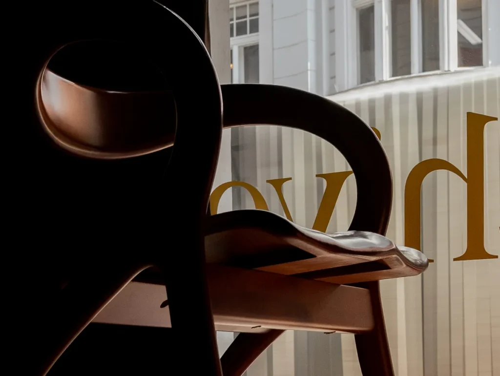 Close-up of a sculptural wooden chair with curved armrest and leather seat, set against a softly lit window with exterior architectural details visible