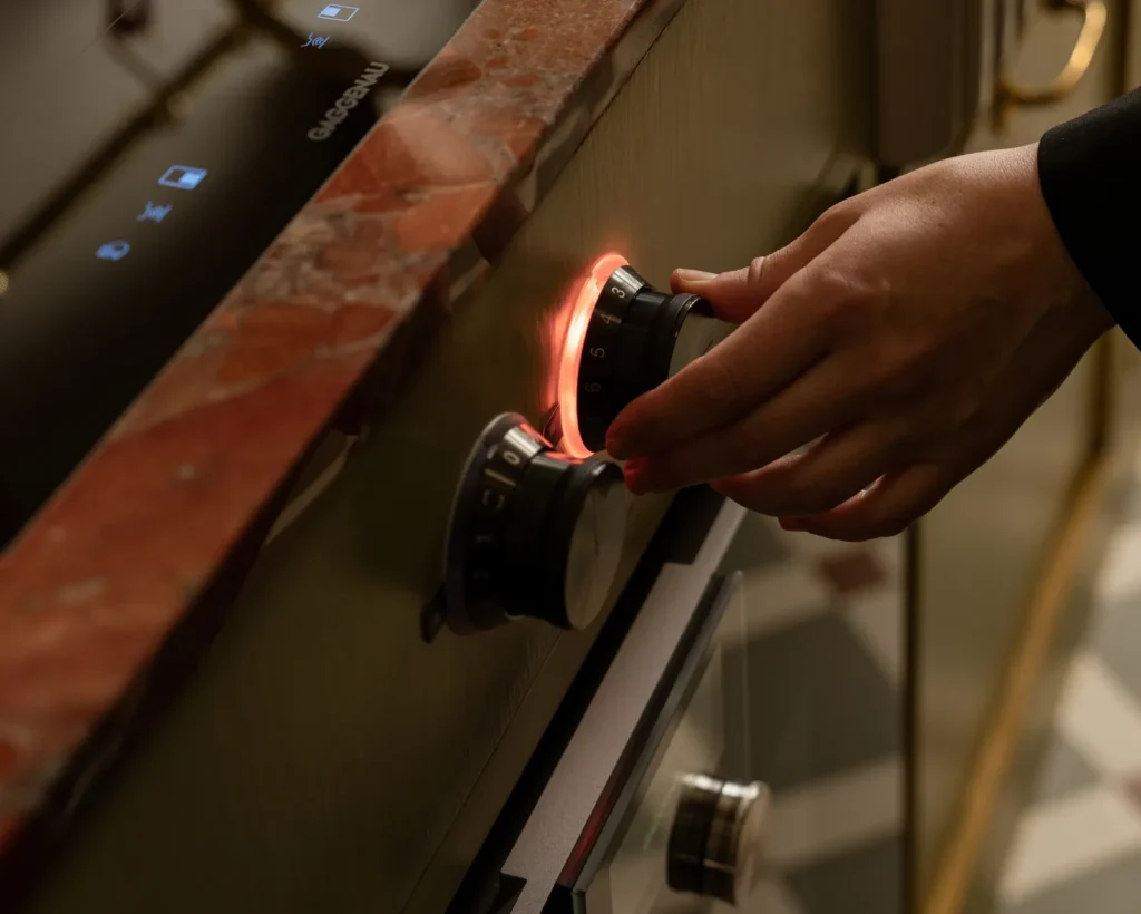 Close-up of a hand adjusting a Gaggenau oven control dial with illuminated ring, set against a marble countertop and wood cabinetry
