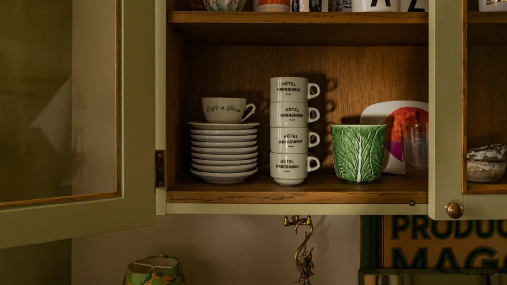 Open kitchen cabinet with neatly stacked cups and saucers, including “Hotel Karasinski” mugs, alongside glassware and decorative ceramics in a warm wood interior