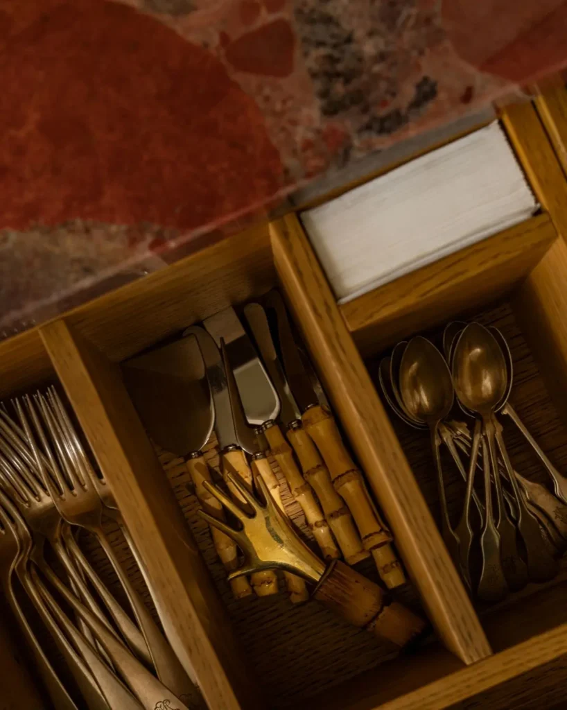 Organized wooden drawer with vintage cutlery, including forks, spoons, and bamboo-handled utensils, beneath a stone countertop in a warm-toned kitchen setting