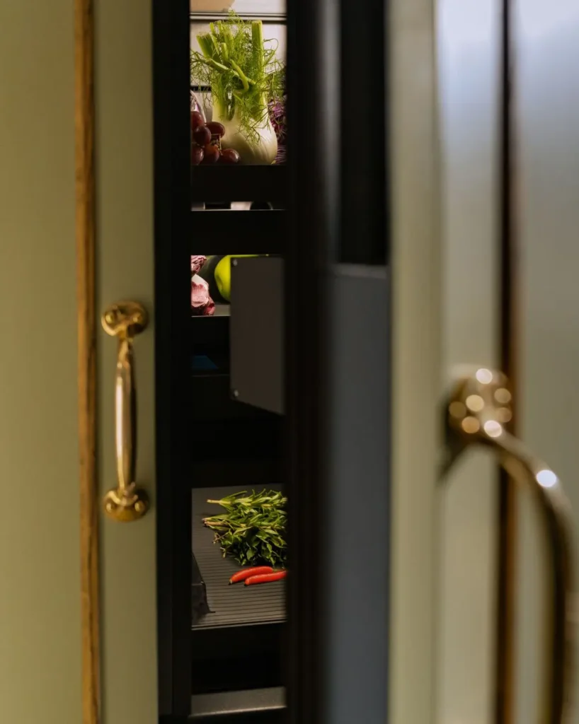 Partially opened integrated refrigerator revealing fresh produce, herbs, and vegetables inside a softly lit, modern kitchen interior