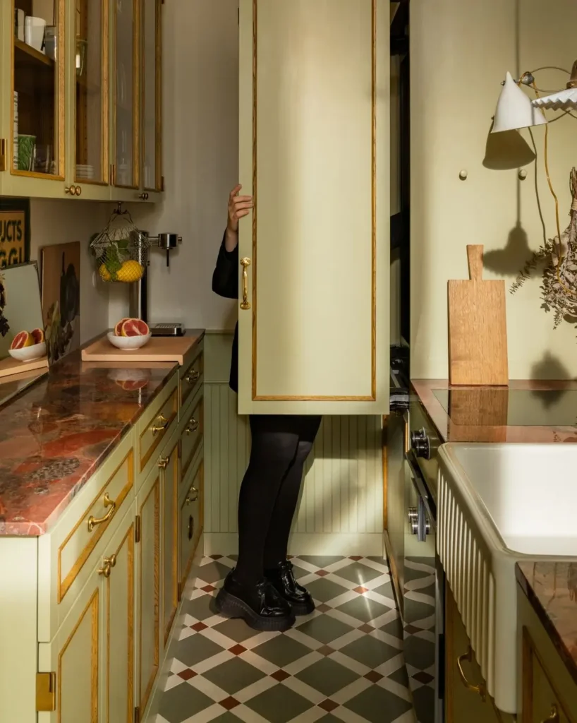 Narrow kitchen with green cabinetry, marble countertops, and patterned tile flooring, as a partially hidden figure opens a tall cabinet in warm natural light