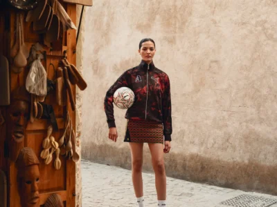 A fashion editorial image set in a narrow, sunlit alley with textured beige walls and a cobblestone ground. A model stands centered, holding a patterned football at her hip, wearing a black zip-up track jacket with red abstract graphics and a matching mini skirt featuring a wavy red-and-green motif. She pairs the look with white socks and vivid green sneakers, creating a sharp contrast against the muted surroundings. To the left, a wooden display of carved masks and hanging utensils adds a handcrafted, artisanal backdrop, reinforcing a dialogue between sport, streetwear, and local craft aesthetics