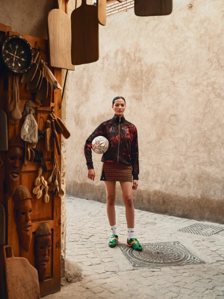A fashion editorial image set in a narrow, sunlit alley with textured beige walls and a cobblestone ground. A model stands centered, holding a patterned football at her hip, wearing a black zip-up track jacket with red abstract graphics and a matching mini skirt featuring a wavy red-and-green motif. She pairs the look with white socks and vivid green sneakers, creating a sharp contrast against the muted surroundings. To the left, a wooden display of carved masks and hanging utensils adds a handcrafted, artisanal backdrop, reinforcing a dialogue between sport, streetwear, and local craft aesthetics