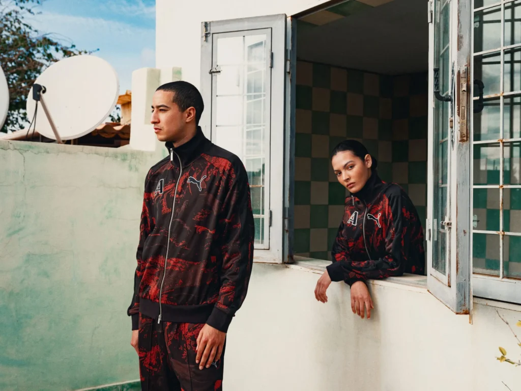 A fashion editorial scene set against a sunlit, slightly weathered building façade with open windows and tiled interiors visible. In the foreground, a model stands in profile wearing a black track jacket and matching pants with red abstract patterns, accented by a logo on the chest. Nearby, another model leans casually out of an open window, dressed in the same coordinated set, gazing directly toward the camera. The setting—complete with a satellite dish and textured walls—adds a lived-in, urban atmosphere that contrasts with the sleek, graphic sportswear aesthetic