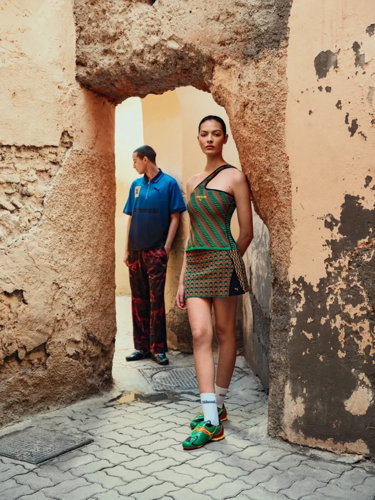 A fashion editorial scene set within a narrow, weathered stone passage framed by a rough archway. In the foreground, a model walks forward wearing an asymmetrical one-shoulder top and matching mini skirt in green and red with a wavy knit pattern, paired with white socks and multicolored sneakers. Behind her, another model stands partially turned away, dressed in a blue “Ahluwalia” jersey-style top with a Puma logo and loose black pants with red abstract detailing. The textured walls and aged surfaces contrast with the bold, graphic garments, creating a cinematic, layered composition