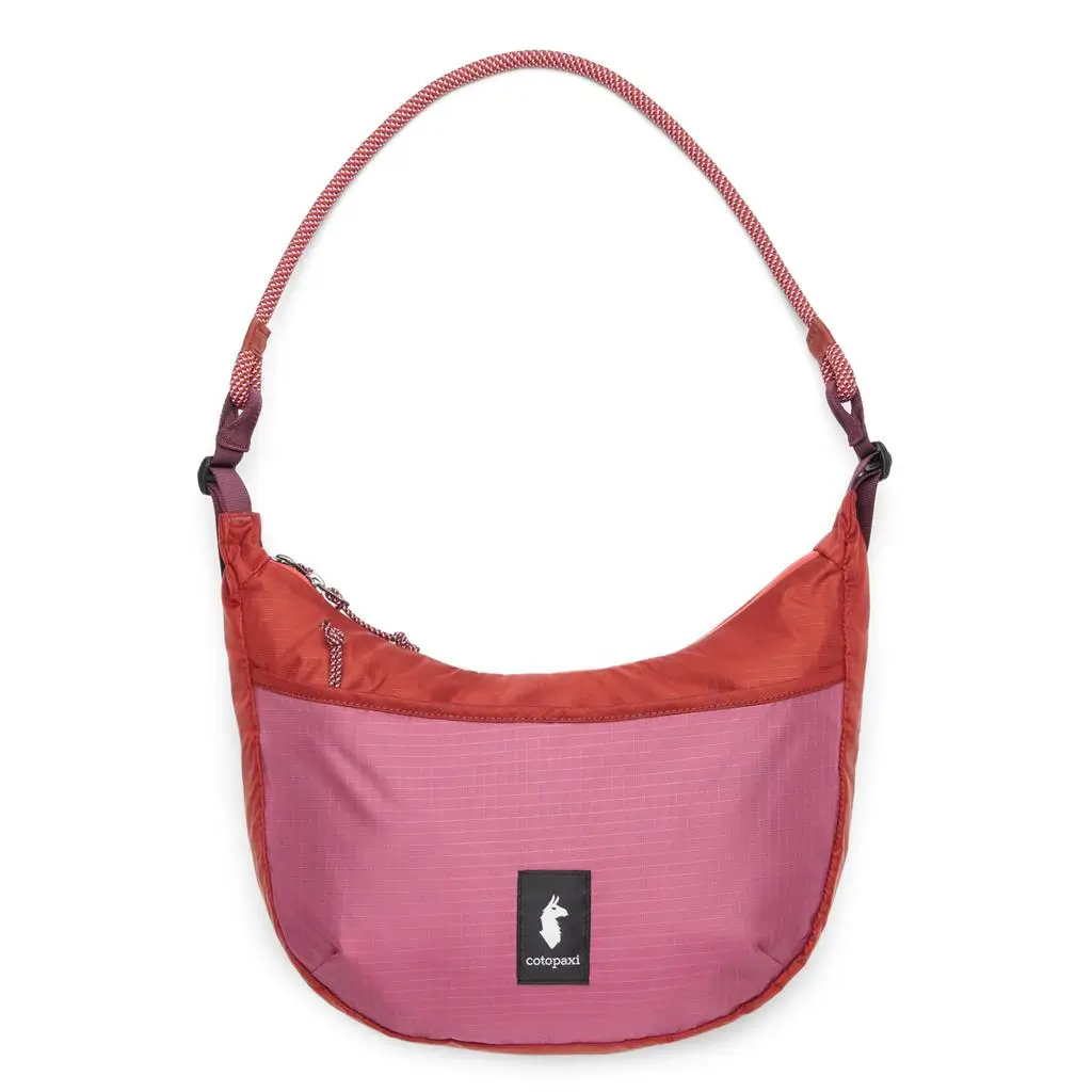 Color-blocked Cotopaxi shoulder bag in deep coral and rose pink ripstop fabric, featuring a curved silhouette, black front logo patch, and a braided adjustable strap with tonal hardware