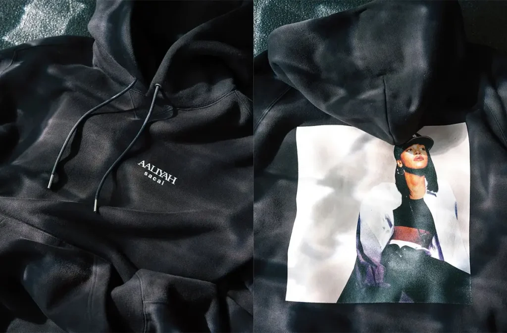 Black sacai hoodie featuring minimalist “AALIYAH sacai” chest logo on one side and a printed portrait of Aaliyah in a cap on the other, set against softly lit textured fabric backdrop