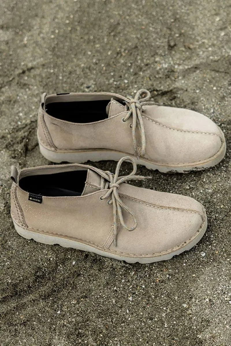 Pair of beige suede moc-toe lace-up shoes with a minimalist Desert Trek–inspired silhouette, featuring tonal stitching, rounded toes, simple eyelets, and lightweight off-white soles, shown on a rough concrete surface