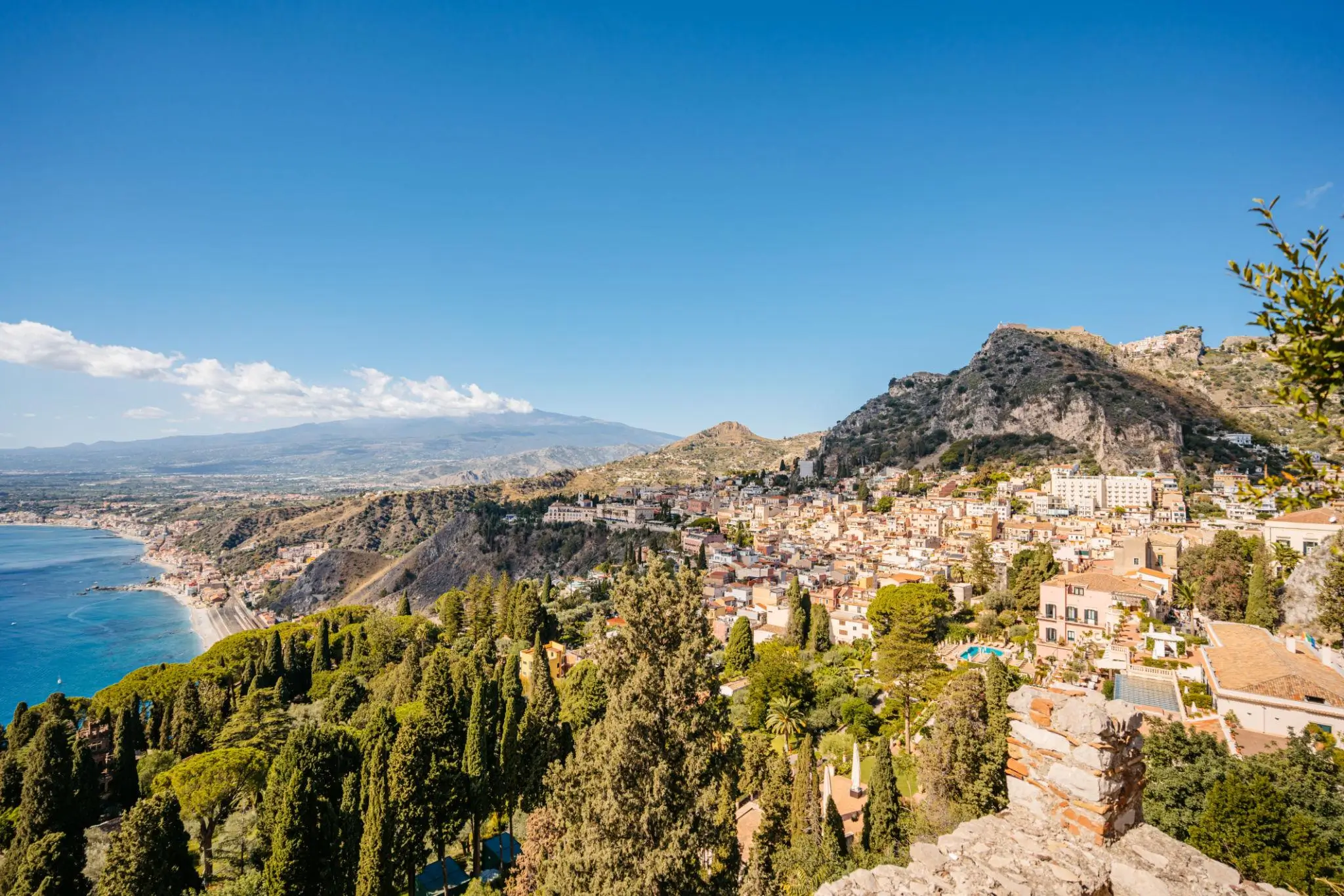 A panoramic view of Taormina, Sicily, with terracotta-roofed buildings cascading down a hillside above the Ionian Sea, framed by lush greenery and cypress trees, while Mount Etna rises in the distance under a clear blue sky