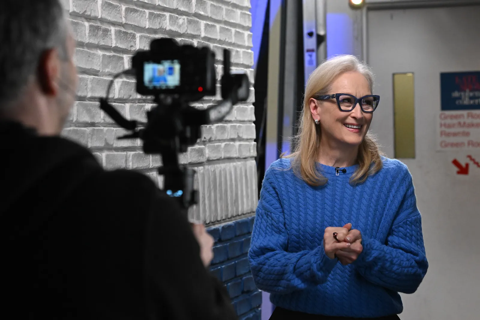 Meryl Streep wearing blue-framed glasses stands indoors against a light gray brick wall, wearing a textured blue sweater. She appears mid-conversation, hands gently clasped, as a camera on a tripod records her from the foreground, suggesting a filmed interview or behind-the-scenes moment in a studio or hallway setting