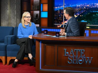 Meryl Streep wearing a blue knit sweater and black trousers sits smiling on a talk show couch, wearing blue-framed glasses, while a male host in a suit speaks to her across a wooden desk on a brightly lit late-night TV set with a city skyline backdrop