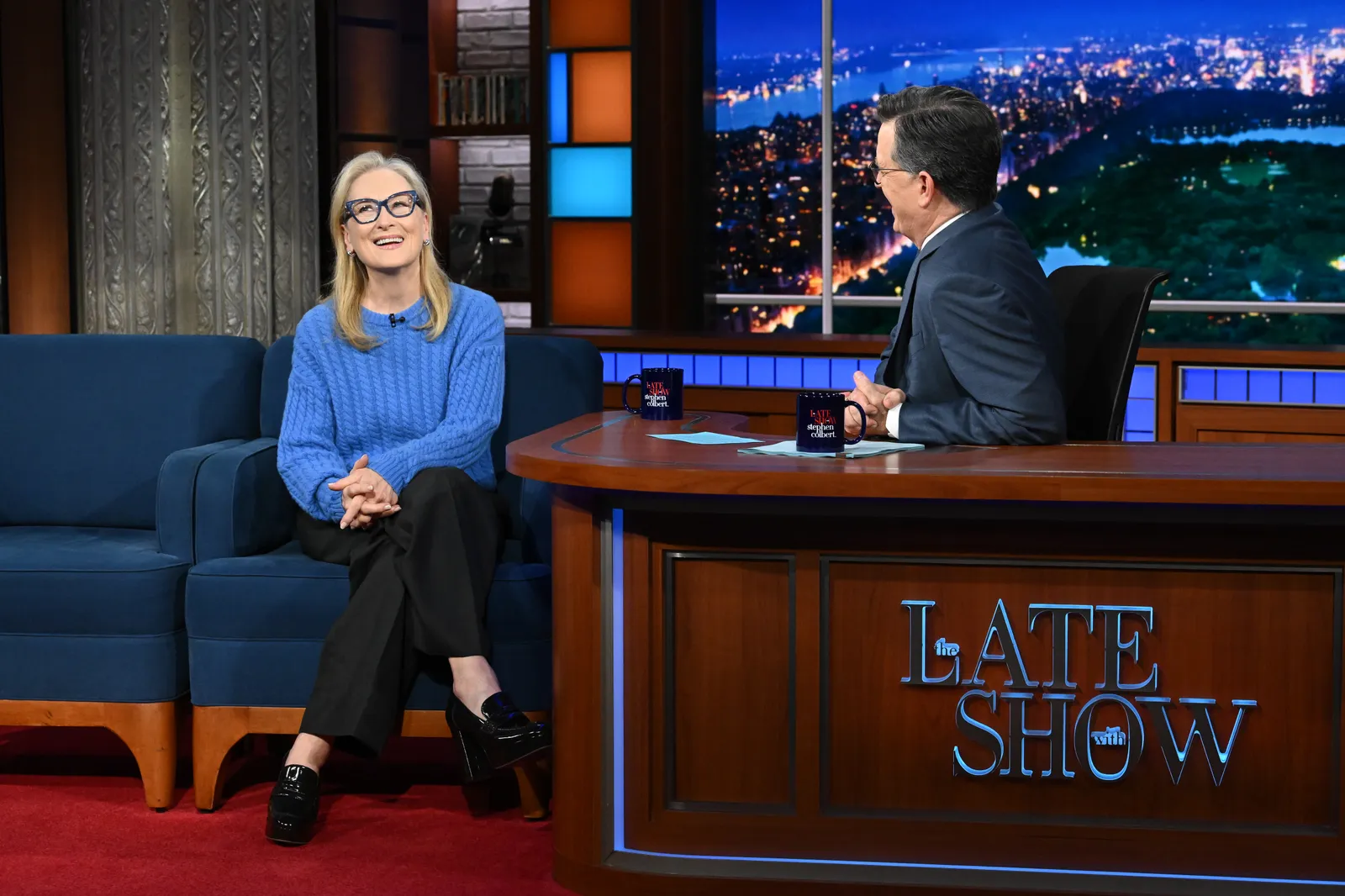 Meryl Streep in a blue knit sweater and blue-framed glasses sits on a talk show couch, hands clasped and looking upward, while a suited Colbert host turns toward her from behind a desk on a brightly lit late-night TV set with a city skyline backdrop